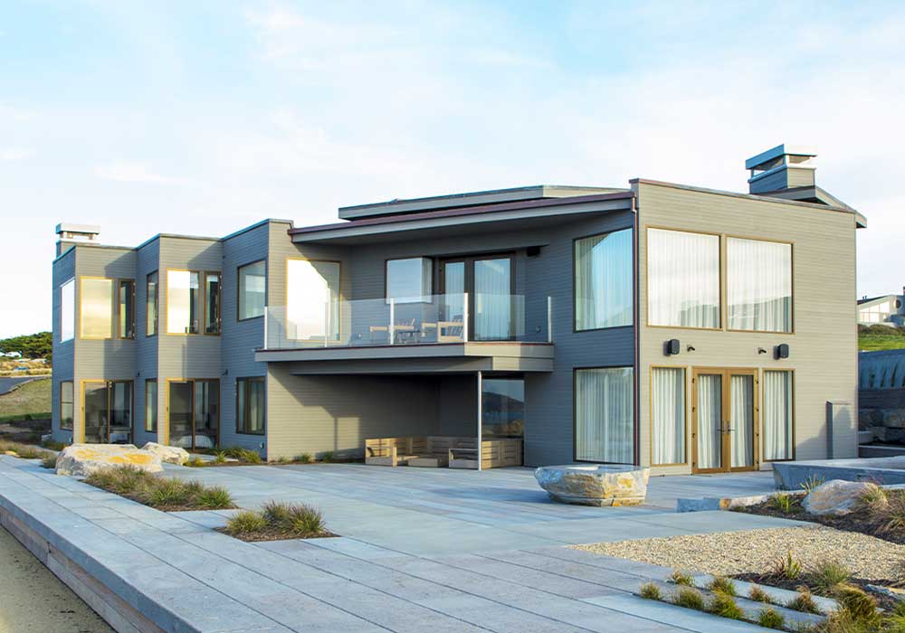 Modern multi-story residential building with large windows and multiple balconies, situated in a coastal area with a clear sky above.